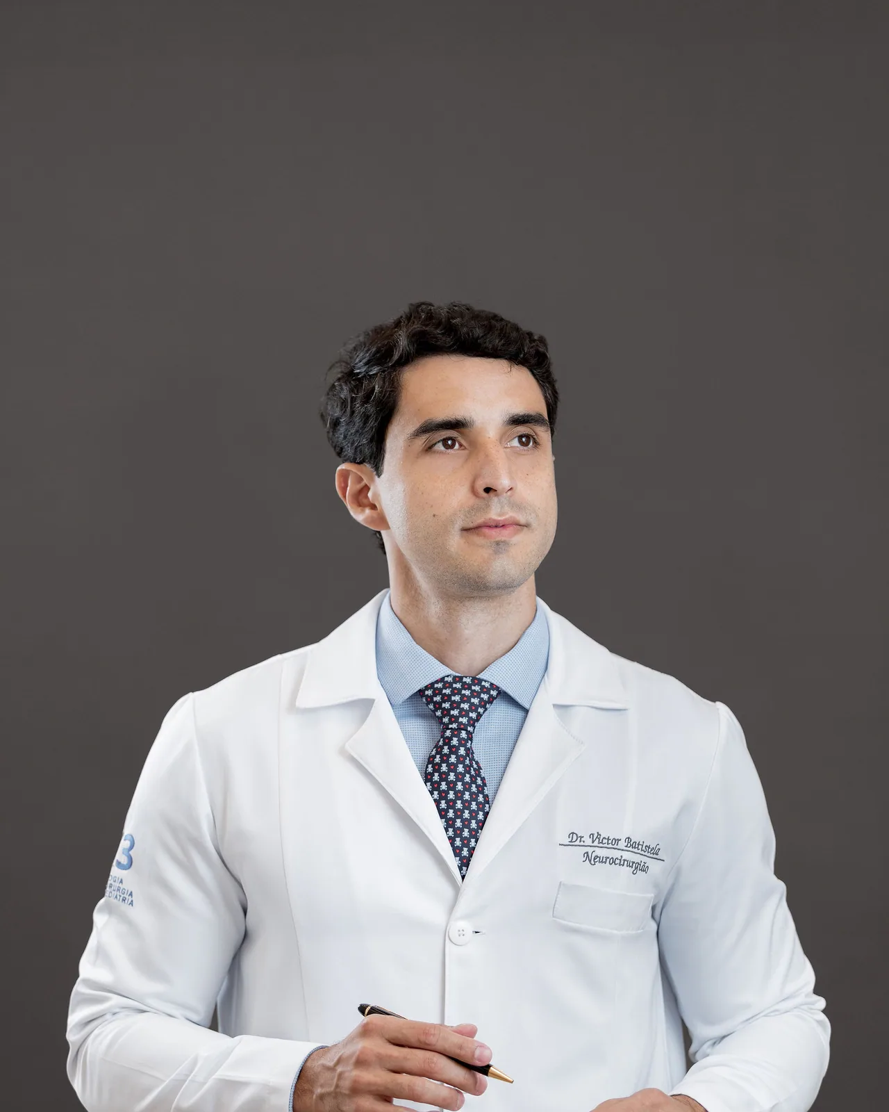 Dr. Victor Batistela de jaleco, com uma caneta na mão, olhando para o lado